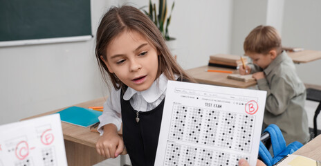 Cute schoolgirl receiving answer sheet with B grade from teacher in classroom