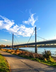 Fototapeta premium A pathway leads to a cable-stayed bridge over water, with a blue sky featuring wispy clouds in the background