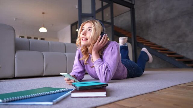 Portrait of joyful cute adolescent girl in wireless headphones relaxing on floor, enjoying music and singing along to catching song while spending leisure in domestic room.