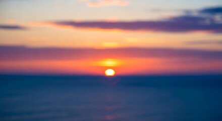 A blurred seascape at sunset showing a bright orange sun over dark water with the sky transitioning through vibrant orange pink and purple tones