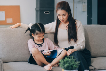 Obraz premium A cheerful Asian mother helps her young daughter do homework and color a book at home, while the father works on a laptop in the background. A cozy, bright, and modern family lifestyle.
