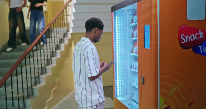 African young adult gen z male man select fast easy choose juice drink press button self service campus place. Happy pupil boy mobile phone use app look screen break time enjoy day indoor college area