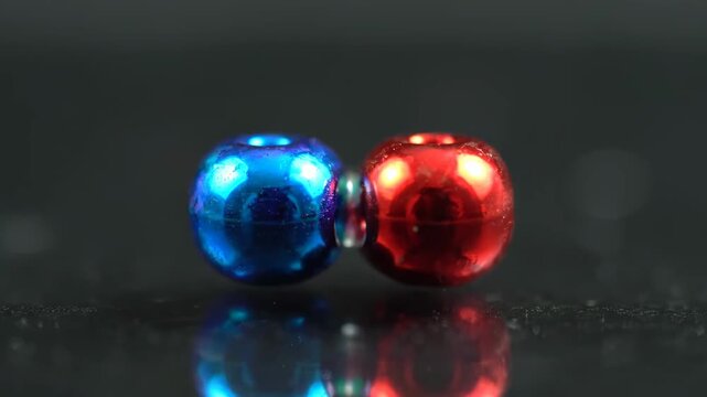 Two vibrant metallic beads, one blue and one red, demonstrate magnetic repulsion on a dark reflective surface. They touch, then forcefully separate.