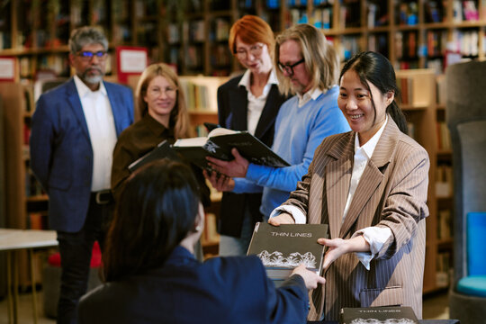 Asian young adult woman handing book to seated female author while diverse group of middle aged men and women standing in background, waiting in line in library during presentation event