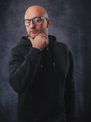 Fototapeta premium A man wearing a black hoodie and glasses is smiling. He is standing in front of a grey background. Classic portrait of a confident and friendly looking male model.