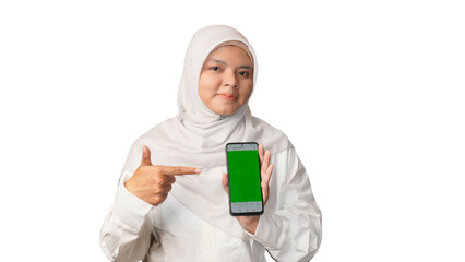 An Asian Indonesian Muslim woman wearing a white dress and headscarf is posing smiling while holding a cellphone and pointing at the green cellphone screen with a white model photo background.