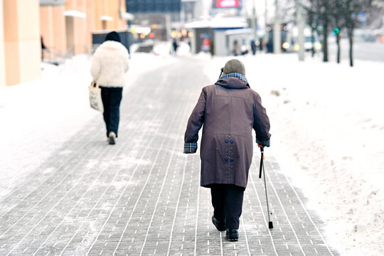 Lone elderly person in long coat and boots walking with support cane on slushy city path covered in fresh snow. Female senior walking with cane on snowy slippery sidewalk during snowfall