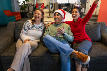 Diverse coworkers laughing on dark gray sofa at office lounge with Santa hat antlers and sweaters