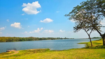 Naklejka premium Panoramic View of a Calm Reservoir Landscape with Blue Sky, Green Grass, and Lush Trees.