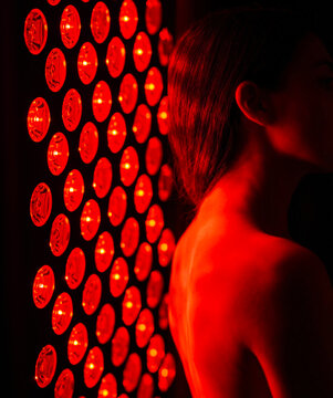 A healthy young woman stands in front of a red light therapy lamp.