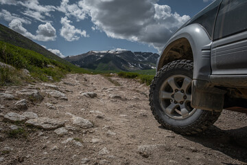 Fototapeta premium Close up of off-road SUV wheel with terrain tire driving on rocky mountain road against snow-capped peaks and blue cloudy sky during summer adventure.