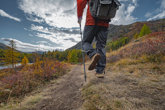 Low angle view of hiker with backpack and trekking poles walking on mountain trail through autumn forest under cloudy sky during wilderness journey.