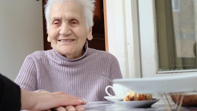 Female hand gently strokes hand of elderly woman sitting at table. Love and care for aging parents, guardianship of seniors and homeless elderly. Sweet gray haired grandmother with bright eyes.