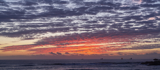 Obraz premium Blood-red sunset over the ocean coast of Portugal with large ships (tankers) visible on the horizon.