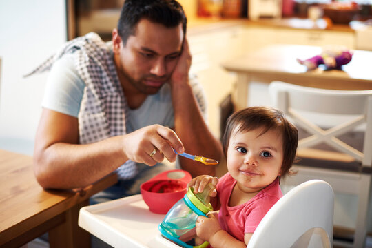Portrait of baby girl sitting in high chair at home with annoyed father in background