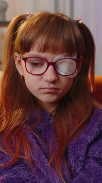 Sick ill preteen girl looking at camera, having protective bandage plaster patch scratch on one eye. Injury, trauma, blind. Child kid wearing glasses at home in room. Medical eyesight vision therapy.
