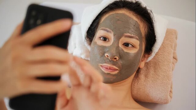 High angle shot of relaxed young Asian woman with applied clay face mask lying on stack of towels and looking at smartphone screen while resting in spa or at home