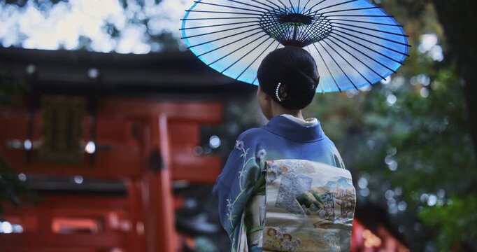 Woman in Japanese kimono holding an umbrella and walking towards torii gate in slow motion