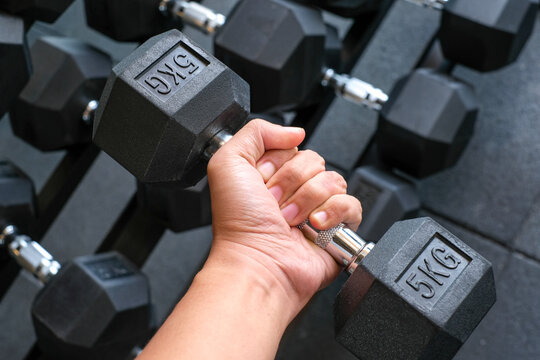 Hand holding a 5kg hex dumbbell with a dumbbell rack in the background