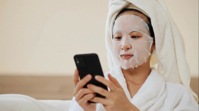 Close up shot of young Asian woman in bathrobe with sheet facial mask using smartphone, scrolling newsfeed or texting friends while enjoying spa day at home