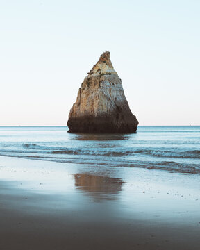 View of a solitary rock formation standing defiant against the gentle waves, its rugged texture mirrored in the wet sand, creating a serene coastal scene, Alvor, Faro, Portugal.