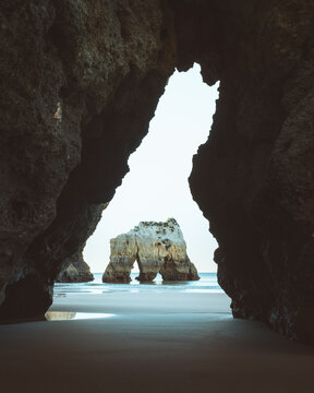 View of the sea arch standing proudly against the horizon, framed by the dark, jagged mouth of a cave on a sandy beach, Alvor, Faro, Portugal.