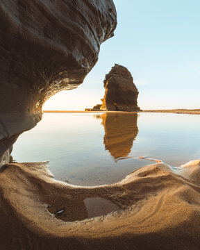 View of a serene tidal pool reflecting a majestic sea stack against a clear sky, framed by rugged rock formations, Vila do Bispo, Faro District, Portugal.