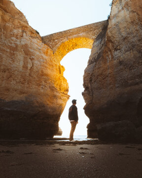 View of a lone figure standing beneath a stone bridge archway, framed by towering cliffs in the golden light, Lagos, Faro, Portugal.