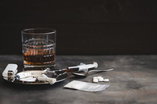 Concept of substance abuse and drug addiction, glass of alcohol, ashtray with cigarettes, lighter, white pills, plastic bag with powder, spoon and syringe with dose on dark background