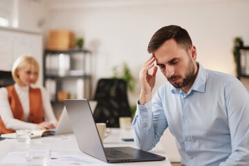 Fototapeta premium A man shows signs of stress as he works on a laptop in an office environment. He holds his head with one hand while another person works in the background.