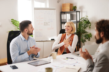 Fototapeta premium In a bright office, a business team gathers around a table. They share ideas and collaborate, expressing support with smiles and clapping. They focus on teamwork and planning.