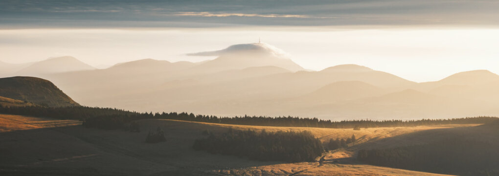 View of golden sunlight kissing the rolling hills and distant, mist-shrouded mountains crowned with a cloud-veiled peak, Mont-Dore, Auvergne-Rhone-Alpes, France.