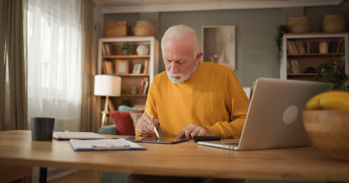 An older man sits at a wooden desk in his living room. He focuses on filling out life insurance paperwork while using a tablet and has a laptop nearby. A banana sits on the table.