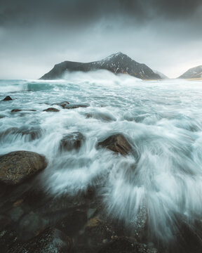 View of turbulent waves crash over dark rocks along the shore, contrasting with the distant, snow-dusted mountain under a brooding sky, Ramberg, Nordland, Norway.