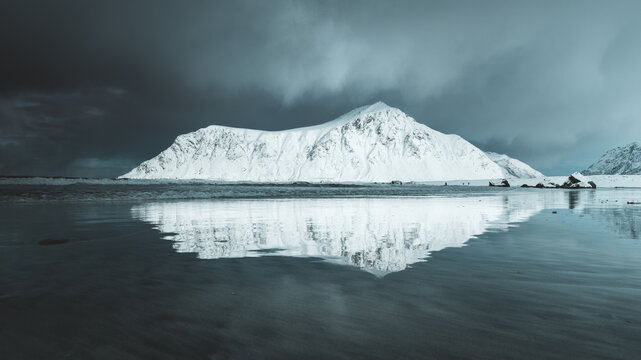 View of a snow-covered mountain mirrored in the calm, icy waters under a moody sky, creating a serene yet dramatic landscape, Ramberg, Nordland, Norway.