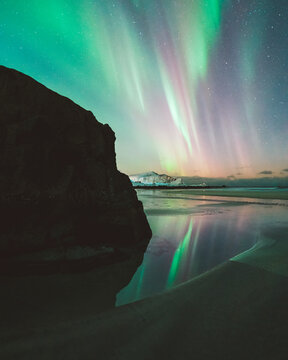 View of vibrant green and pink aurora borealis dancing above the still, reflective waters and dark rocks, creating a magical scene, Ramberg, Nordland, Norway.
