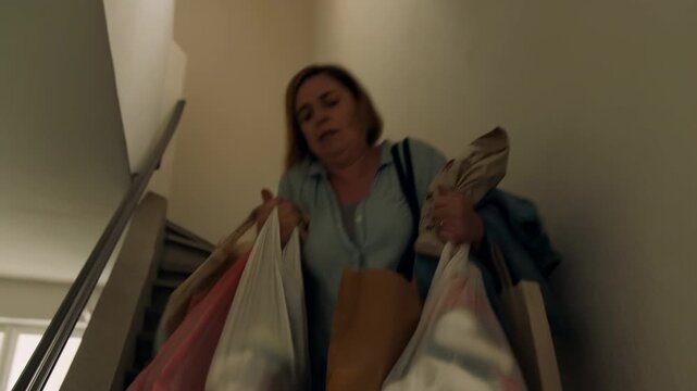 An Exhausted Woman Carefully Carries Several Grocery Bags Down Confined Apartment Staircase After Shopping