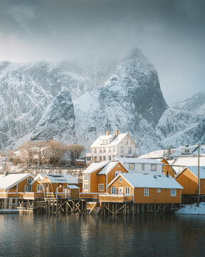 View of golden cabins meet the icy waters beneath snow-laden peaks, a serene blend of warmth and winter's chill in Reine, Nordland, Norway.
