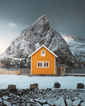 View of a vibrant yellow house contrasts against a monumental snow-clad mountain under a moody sky, creating a striking winter scene, Reine, Nordland, Norway.
