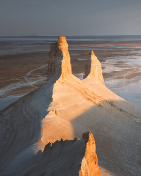 View of towering rock formations kissed by the sun's golden light, contrasting with the vast, flat landscape, creating a surreal scene, Aktau, Kazakhstan.
