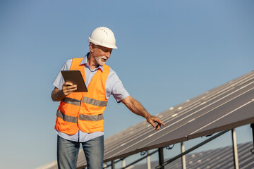 Senior white male environmentalist in safety gear inspecting solar panels at solar farm during clear day