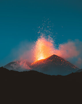 View of volcano erupting, spewing fiery lava and ash against the twilight sky, the dark silhouette of the surrounding landscape, Nicolosi, Sicily, Italy.