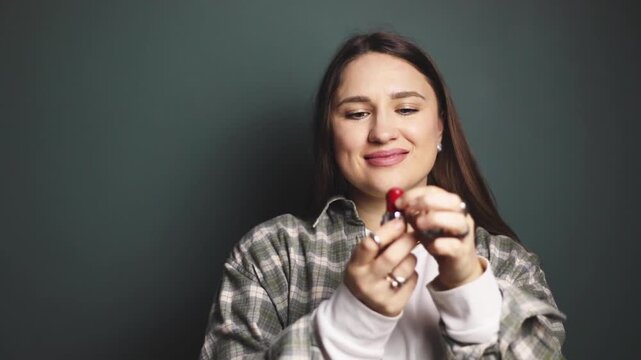 Smiling brunette woman applying bright blush stick on cheekbones and blending with finger, natural beauty makeup routine, female model demonstrating cosmetic product on gray green background, studio 