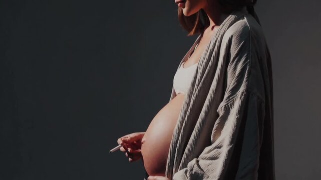 A side-lit woman in a soft robe and bralette holds a lit cigarette against a dark gray backdrop; warm highlights trace the curve of her rounded belly as smoke curls upward in a quiet, tense moment.