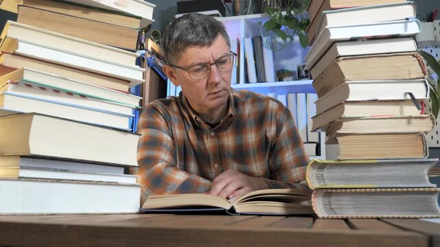 Focused man reading between tall stacks of books at home desk, Serious self education and research in quiet indoor workspace environment