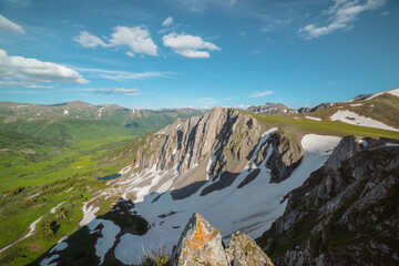 Aerial dizzying view to rocky cliff and sharp rocks under clouds in blue sky. Vertigo scenery with sheer crags above small lake in green dale and mountain top far away in cloudy changeable weather. © Daniil