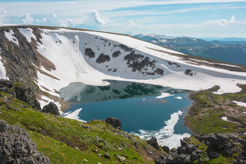 Scenic sunlit landscape with alpine lake in rocky snowy cirque near stone hill top during thaw. Ice floats in mountain lake among rocks and snows with view to forest mountain range under cloudy sky. © Daniil