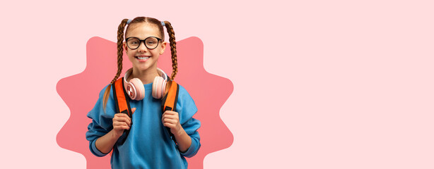 A young girl stands with a backpack and headphones around her neck. She has glasses and a joyful expression. The background is a simple pink color, making her look bright and cheerful. © Prostock-studio