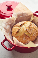 Golden sourdough bread with cracked crust in red ceramic Dutch oven, lined with parchment paper. Perfect for culinary, baking, and food photography projects.