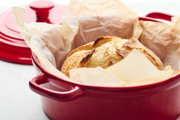 Golden sourdough bread with cracked crust in red ceramic Dutch oven, lined with parchment paper. Perfect for culinary, baking, and food photography projects.